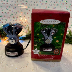 May include: A Harley-Davidson Big Twin Evolution engine ornament. The ornament is silver and black, with the Harley-Davidson logo on the base. The box reads "Keepsake Ornament" and "Lights and Motion".