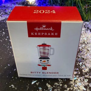 Puede incluir: Caja de adorno navideño Hallmark. La caja es blanca con la parte superior roja y presenta un diseño de licuadora. La licuadora es blanca y roja, con una cara de muñeco de nieve en la base. El texto "2024" está en la parte superior.