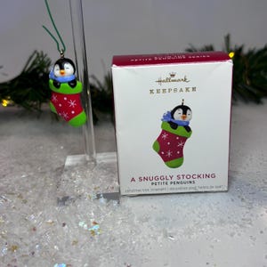 May include: A Christmas tree ornament featuring a penguin in a red and green stocking. The penguin has a blue scarf and is smiling. The ornament is hanging in front of a box with the text "A Snuggly Stocking Petite Penguins".