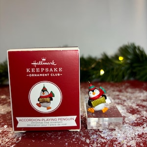 May include: A Hallmark Keepsake Christmas tree ornament featuring an accordion-playing penguin. The penguin is wearing a red scarf and a green hat. The ornament is on a clear display stand, with the red box in the background.