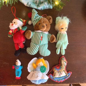 May include: A collection of six vintage Christmas ornaments. The ornaments include a Santa Claus, a teddy bear wearing pajamas, a rocking horse, an angel, and two dolls wearing pajamas.
