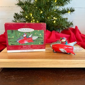 May include: A red and silver Christmas ornament shaped like a rocket sled with Santa Claus at the helm. The ornament is labeled "Sleigh X-2000" and comes with its original box, which also features the ornament's image.