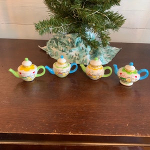 May include: Four miniature porcelain teapots with lids. Each teapot is painted with a different design featuring teacups, teapots, and tea-related imagery. The teapots are in shades of yellow, green, blue, and white.