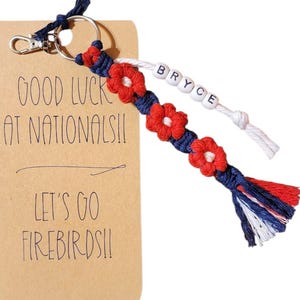 May include: A handmade keychain with a red, white, and blue braided design and red flower accents. The keychain includes a tag with the text "GOOD LUCK AT NATIONALS!! LET'S GO FIREBIRDS!!" and the name "BRYCE" on a separate strand.