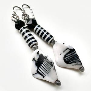 May include: Dangle earrings with a monochrome design. The earrings have silver-tone hooks, black and white striped beads, and white, irregular pendants with black abstract patterns. The earrings are set against a white background.
