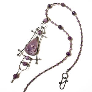 May include: A necklace featuring a teardrop-shaped purple stone pendant with textured circular inclusions, set in a dark metal frame. The chain consists of alternating small and large purple beads interspersed with thin metal segments and a hook clasp.