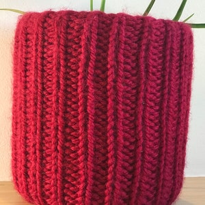 Puede incluir: Una funda de maceta de punto de color rojo oscuro para una planta. La funda está hecha de hilo grueso y tiene una textura acanalada.