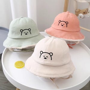 May include: Three children's bucket hats with a bear design. The hats are in green, pink, and cream colours.