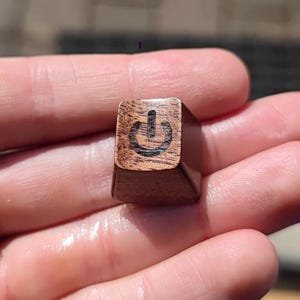 May include: A wooden keyboard key with a power symbol engraved on it. The key is brown and has a slightly textured surface.