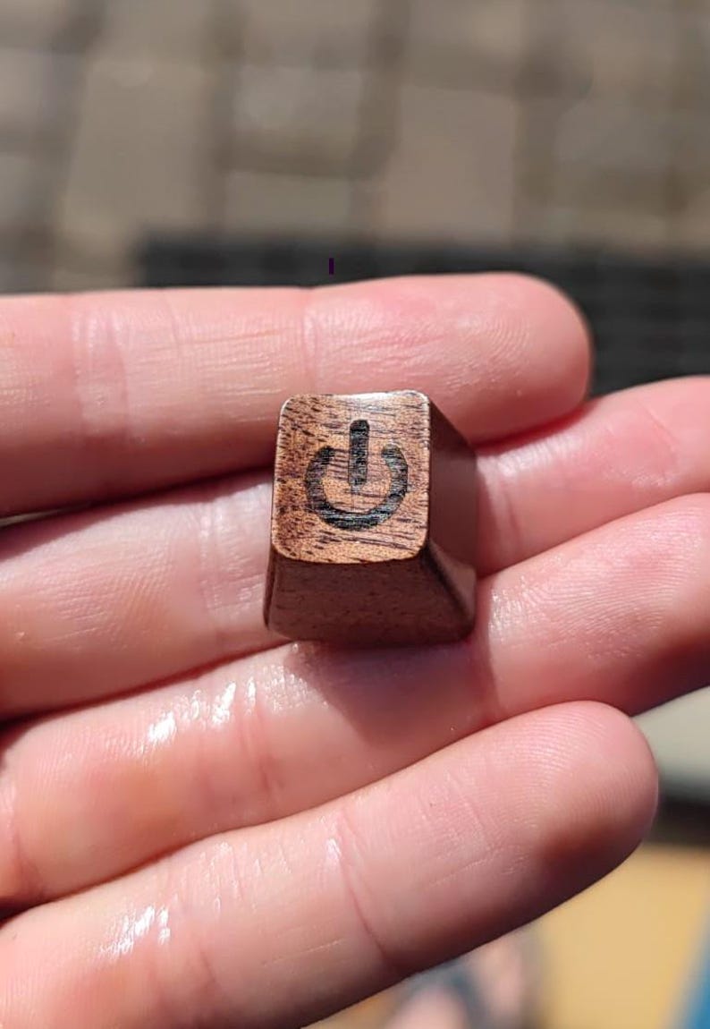May include: A wooden keyboard key with a power symbol engraved on it. The key is brown and has a slightly textured surface.