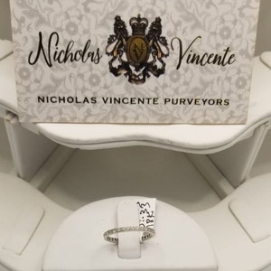 Puede incluir: Una fina banda de oro blanco con peque&ntilde;os diamantes engastados en pav&eacute;. El anillo se muestra en un expositor de anillos blanco. El texto "Nicholas Vincente Purveyors" es visible en una tarjeta blanca detr&aacute;s del anillo.