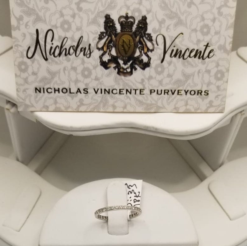 Puede incluir: Una fina banda de oro blanco con peque&ntilde;os diamantes engastados en pav&eacute;. El anillo se muestra en un expositor de anillos blanco. El texto "Nicholas Vincente Purveyors" es visible en una tarjeta blanca detr&aacute;s del anillo.