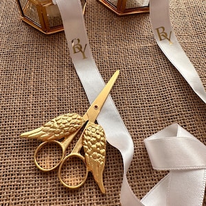 May include: Gold-colored scissors with wing-shaped handles rest on a textured surface, next to a white ribbon with gold lettering. The scissors are the focal point, with a detailed wing design. The ribbon has the letters "BY" printed on it.