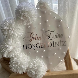 May include: A clear, round sign with the words "Gelin Evine Hoşgeldiniz" in silver script, surrounded by white pom-pom flowers and faux pearls. The sign is displayed on a small wooden easel.