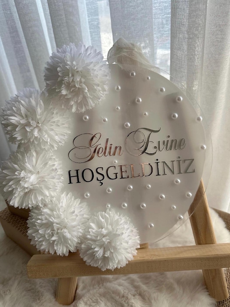 May include: A clear, round sign with the words "Gelin Evine Hoşgeldiniz" in silver script, surrounded by white pom-pom flowers and faux pearls. The sign is displayed on a small wooden easel.