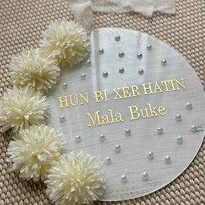 May include: A round, clear acrylic sign with gold lettering that reads "H&Ugrave;N BI X&Egrave;R HATIN Mala Buke". The sign is decorated with white faux flowers and small pearl beads. A white ribbon is attached at the top.