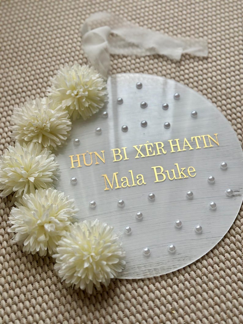 May include: A round, clear acrylic sign with gold lettering that reads "H&Ugrave;N BI X&Egrave;R HATIN Mala Buke". The sign is decorated with white faux flowers and small pearl beads. A white ribbon is attached at the top.