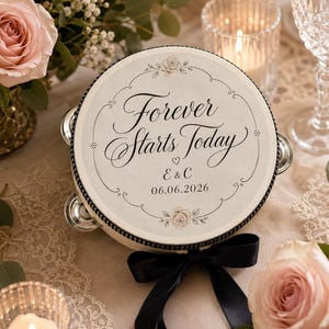 May include: A round tambourine featuring a cream-colored surface with the black text Forever Starts Today, the initials E & C, and the date 06.06.2026. The design includes floral accents and a black ribbon tied at the bottom with metallic jingles.