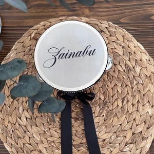 May include: A circular tambourine with a smooth, light-colored drumhead featuring the name Zainabu in elegant black script. The instrument is accented with a black ribbon tied in a bow at the bottom and includes small metal jingles along the rim.