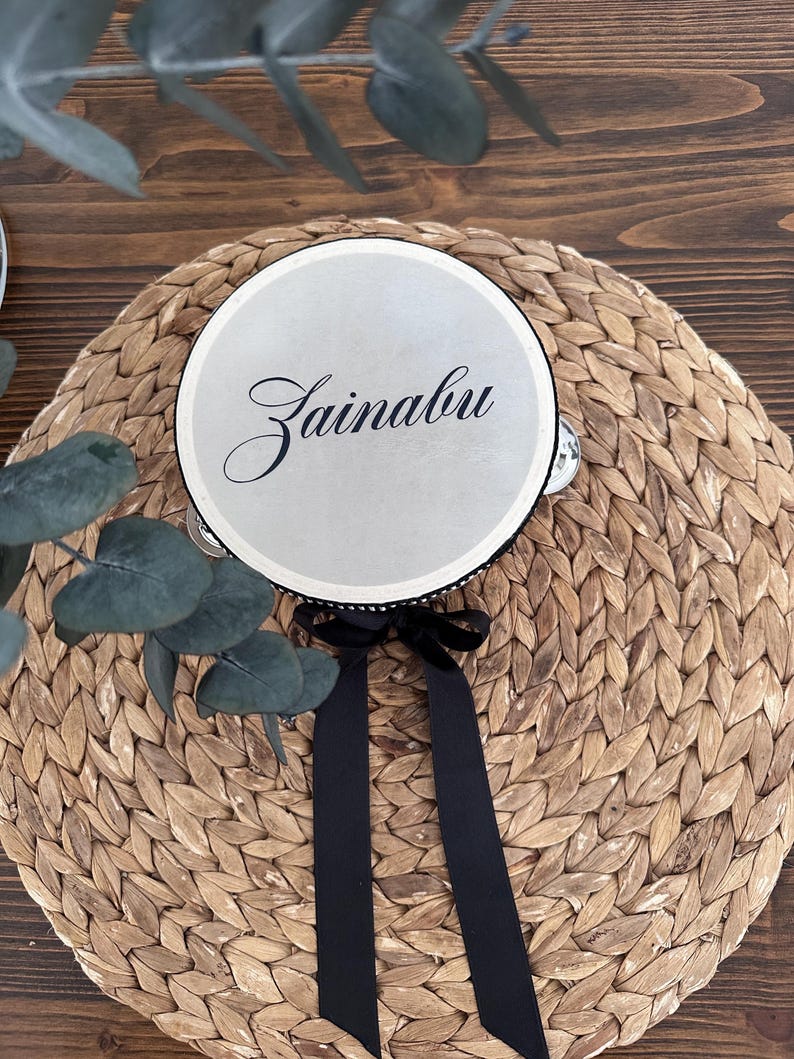 May include: A circular tambourine with a smooth, light-colored drumhead featuring the name Zainabu in elegant black script. The instrument is accented with a black ribbon tied in a bow at the bottom and includes small metal jingles along the rim.
