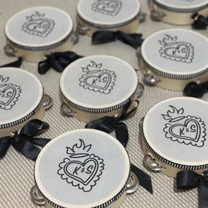 May include: A collection of round tambourines with cream-colored frames and black and white striped trim. Each tambourine features a light gray top printed with a black line-art sacred heart design containing the letters K&S and a black ribbon bow.