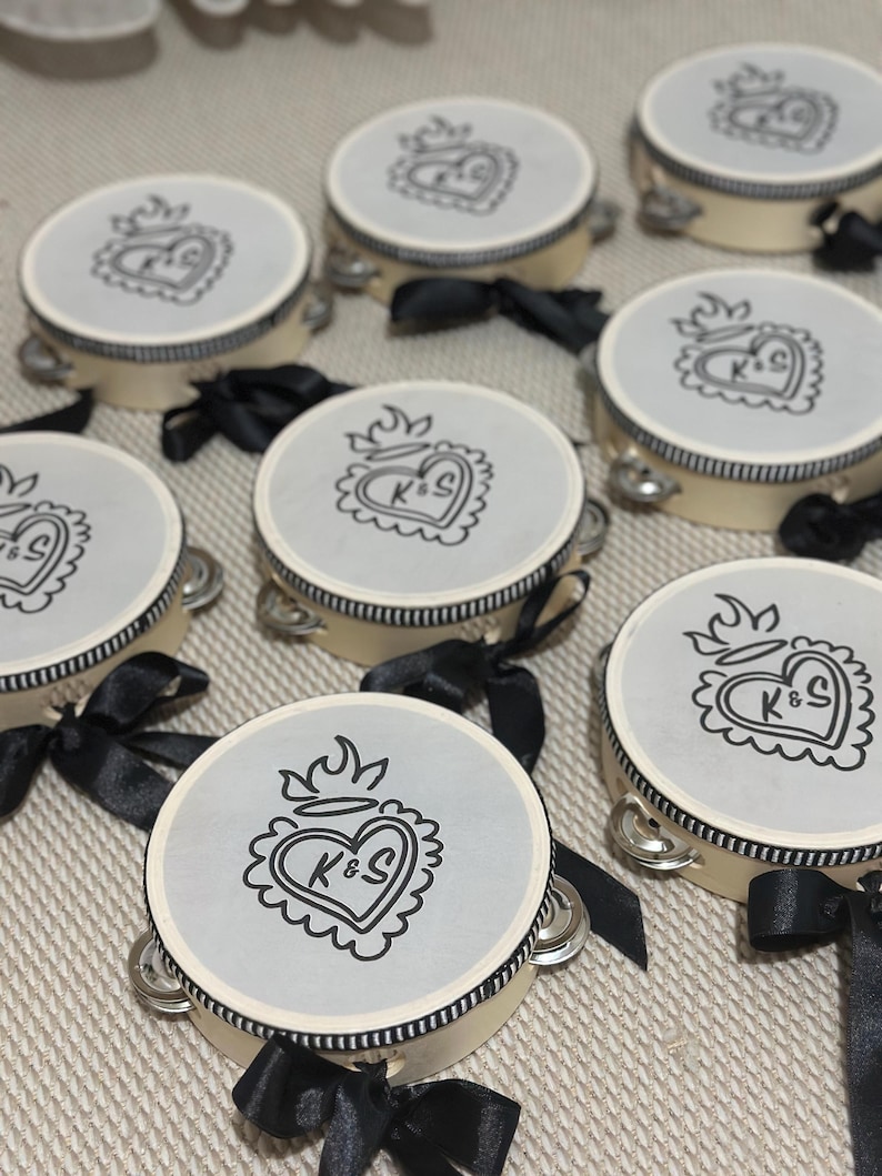 May include: A collection of round tambourines with cream-colored frames and black and white striped trim. Each tambourine features a light gray top printed with a black line-art sacred heart design containing the letters K&S and a black ribbon bow.