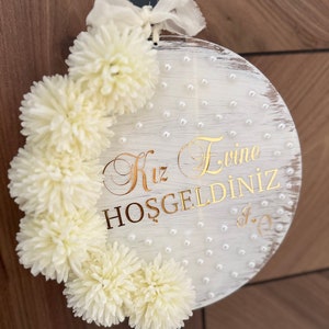 May include: A round, white wooden sign with gold lettering and cream-colored pom-poms. The sign has a distressed finish and is adorned with small, pearl-like beads. The text reads "Kız Evine HOŞGELDİNİZ". A cream-colored ribbon is attached at the top.