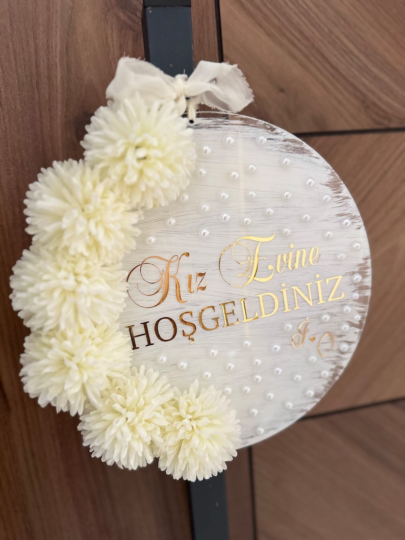 May include: A round, white wooden sign with gold lettering and cream-colored pom-poms. The sign has a distressed finish and is adorned with small, pearl-like beads. The text reads "Kız Evine HOŞGELDİNİZ". A cream-colored ribbon is attached at the top.