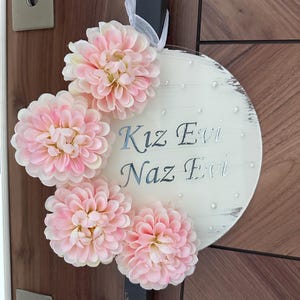 May include: A round, white wooden sign with the text "Kız Evi Naz Evi" in silver script. The sign is decorated with four large, light pink artificial flowers and small pearl accents. A white ribbon is attached for hanging.