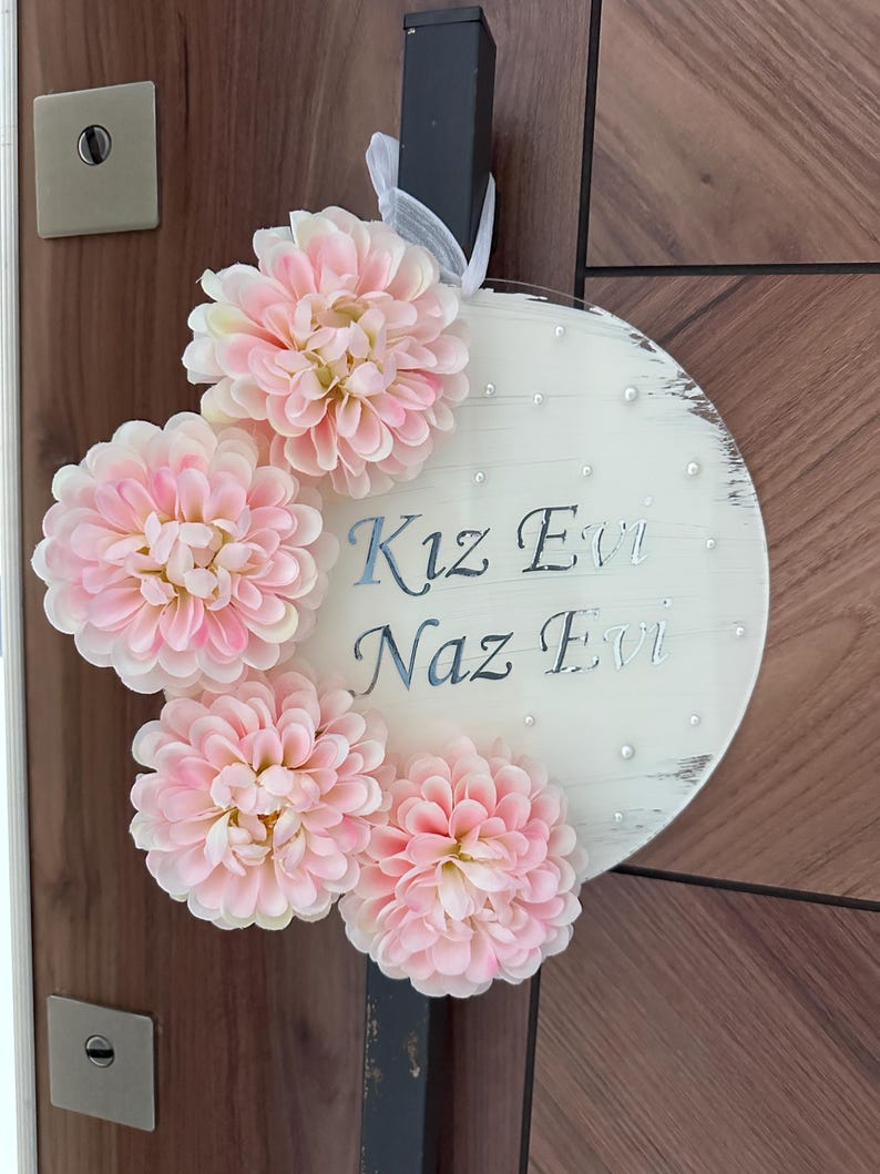 May include: A round, white wooden sign with the text "Kız Evi Naz Evi" in silver script. The sign is decorated with four large, light pink artificial flowers and small pearl accents. A white ribbon is attached for hanging.