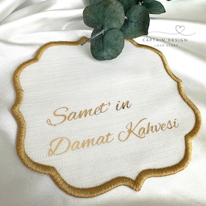 Puede incluir: Mantel individual bordado blanco con ribete dorado y texto dorado que dice "Samet in Damat Kahvesi". El mantel individual tiene un borde festoneado y está decorado con una ramita de hojas verdes.