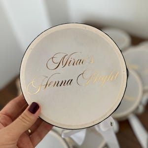 May include: A round, off-white drum-shaped container with a black rim and a white ribbon attached. The top surface features the gold-colored text "Mirae's Henna Night" written in a decorative, elegant script font.