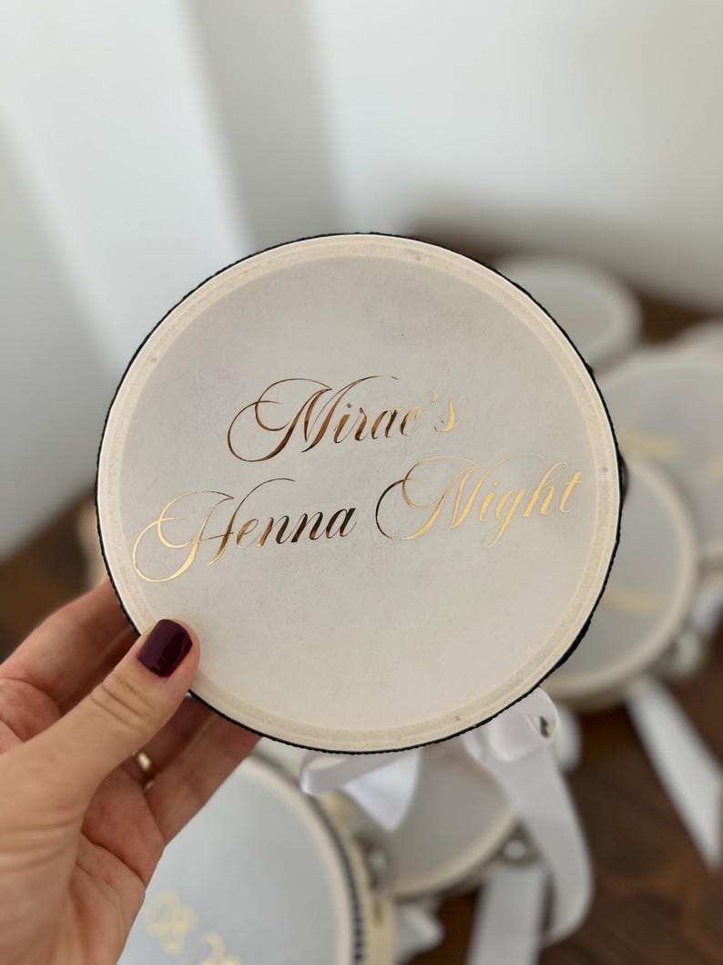May include: A round, off-white drum-shaped container with a black rim and a white ribbon attached. The top surface features the gold-colored text "Mirae's Henna Night" written in a decorative, elegant script font.