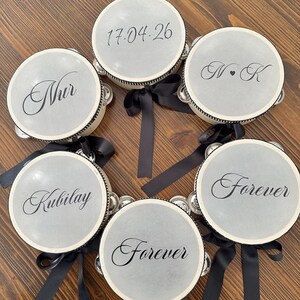 May include: A collection of six circular tambourines with light gray drumheads, each featuring black script text including names, dates, and the word Forever. The frames are white with black trim and each is adorned with a black satin ribbon bow.