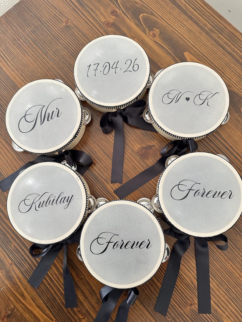 May include: A collection of six circular tambourines with light gray drumheads, each featuring black script text including names, dates, and the word Forever. The frames are white with black trim and each is adorned with a black satin ribbon bow.