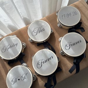 May include: Six round tambourines with light-colored drumheads and black rims. Each features black text: 'Nur', 'Kubilay', 'Forever', '17.04.26', and 'N&bull;K'. Each tambourine is decorated with a black ribbon tied to the side, featuring metal jingles.