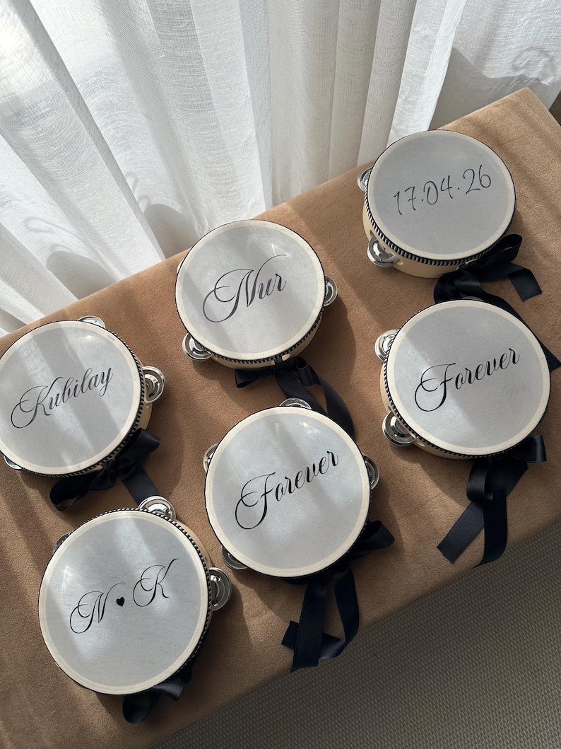 May include: Six round tambourines with light-colored drumheads and black rims. Each features black text: 'Nur', 'Kubilay', 'Forever', '17.04.26', and 'N&bull;K'. Each tambourine is decorated with a black ribbon tied to the side, featuring metal jingles.