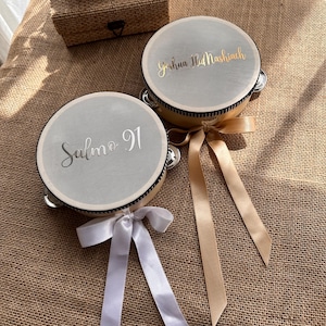 May include: Two round tambourines with light gray drumheads. One features the silver text Salmo 91, the other gold text Yeshua HaMashiach. Each has a wooden frame, metal jingles, and a decorative ribbon in white or gold tied to the side.