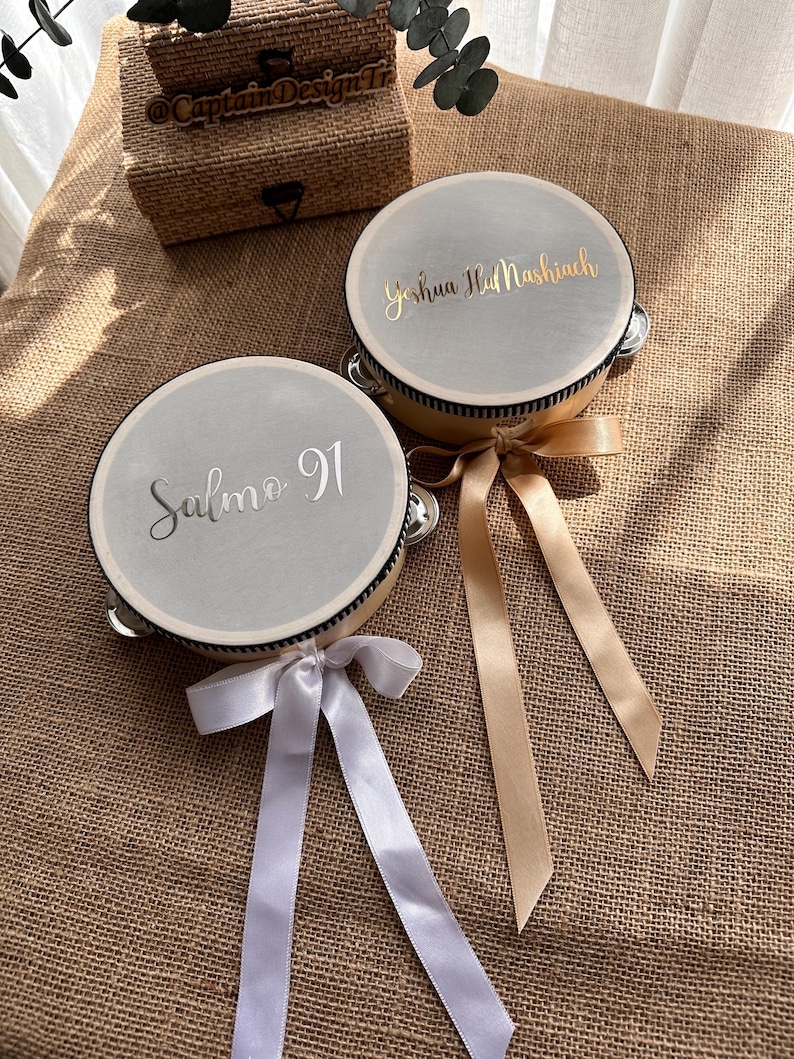 May include: Two round tambourines with light gray drumheads. One features the silver text Salmo 91, the other gold text Yeshua HaMashiach. Each has a wooden frame, metal jingles, and a decorative ribbon in white or gold tied to the side.
