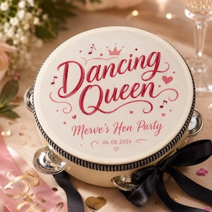 May include: A round tambourine with a cream-colored head featuring the red text Dancing Queen, Merve's Hen Party, and the date 06.08.2026. The design includes a small crown, musical notes, and hearts. A black ribbon is tied to the side frame.