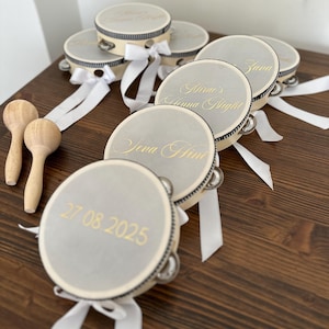 May include: A collection of round tambourines with light gray drumheads, featuring gold-colored text including names, dates, and the phrase Henna Night. Each tambourine has a black and white striped rim, silver jingles, and a white satin ribbon.