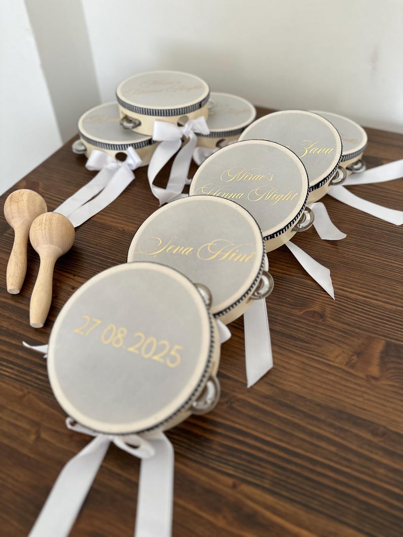 May include: A collection of round tambourines with light gray drumheads, featuring gold-colored text including names, dates, and the phrase Henna Night. Each tambourine has a black and white striped rim, silver jingles, and a white satin ribbon.