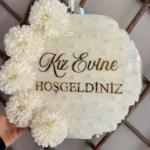 May include: Round, clear acrylic sign with white paint and faux pearls. The sign features the gold text "Kız Evine HOŞGELDİNİZ" and is adorned with white floral accents. A decorative piece for a home.