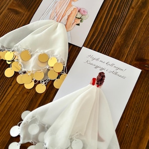 Puede incluir: Dos velos de novia de tela blanca con lentejuelas doradas y blancas. Los velos están doblados y expuestos sobre una superficie de madera. El texto en la tarjeta dice "Hajde me kall nejon! Kanagyezi i Athane's".