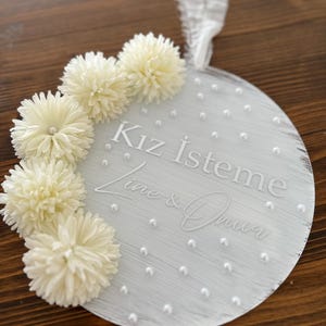 May include: A round, white decorative sign with the text "Kız İsteme" and names. The sign is adorned with white pom-pom flowers and small pearl accents. A white ribbon is attached for hanging.