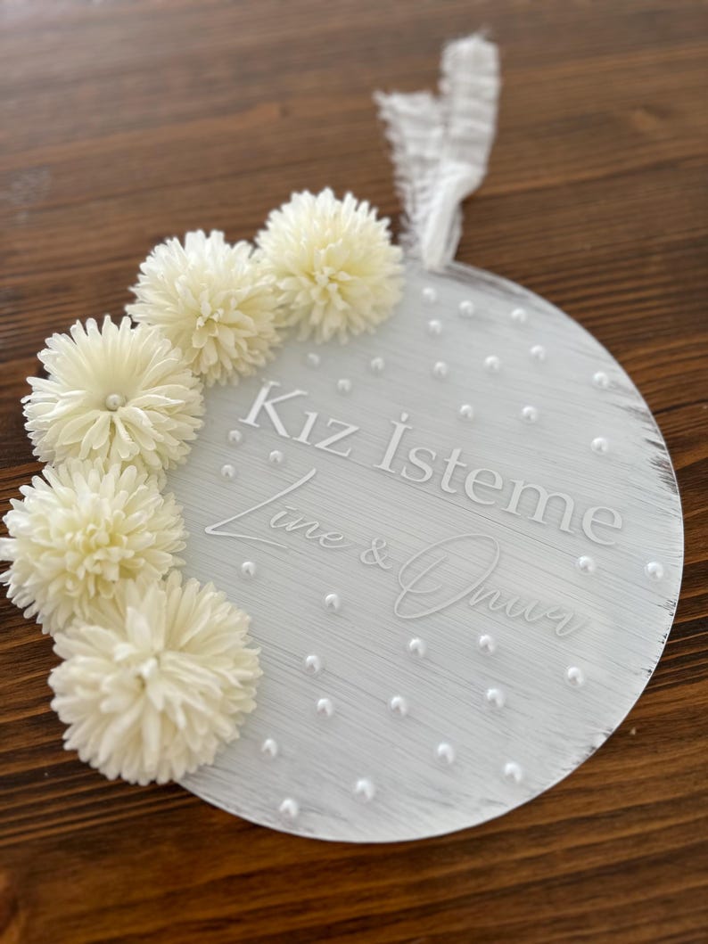 May include: A round, white decorative sign with the text "Kız İsteme" and names. The sign is adorned with white pom-pom flowers and small pearl accents. A white ribbon is attached for hanging.