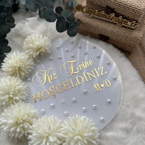 May include: A round, clear acrylic sign with gold lettering that reads "Kız Evine Hoşgeldiniz M&hearts;D". The sign is decorated with small pearls and white flowers. Two woven boxes are in the background.