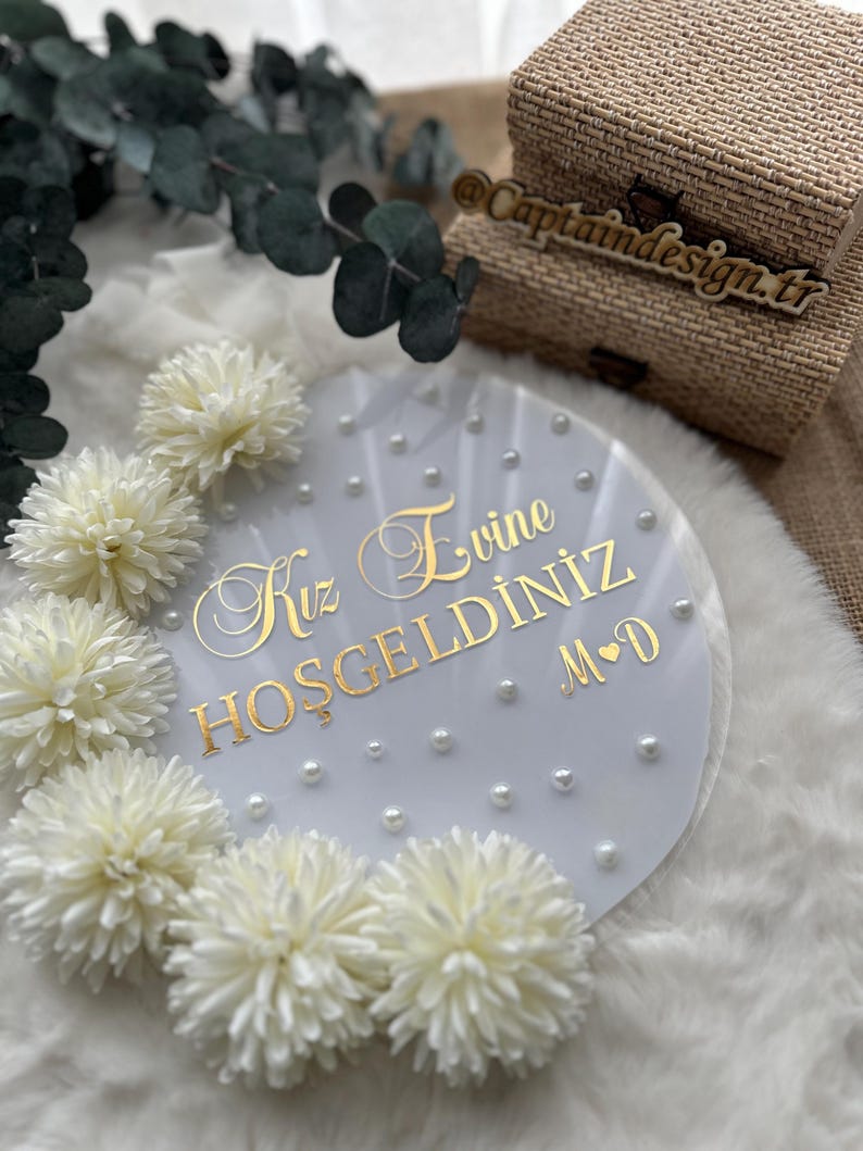 May include: A round, clear acrylic sign with gold lettering that reads "Kız Evine Hoşgeldiniz M&hearts;D". The sign is decorated with small pearls and white flowers. Two woven boxes are in the background.