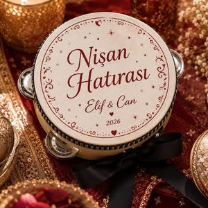 May include: A round tambourine with a cream-colored surface featuring the dark red text "Nişan Hatırası", "Elif & Can", and "2026". The instrument is decorated with a black ribbon bow and small metallic jingles around the circular frame.