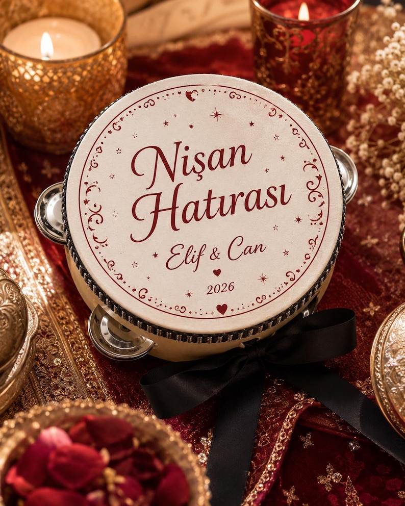 May include: A round tambourine with a cream-colored surface featuring the dark red text "Nişan Hatırası", "Elif & Can", and "2026". The instrument is decorated with a black ribbon bow and small metallic jingles around the circular frame.
